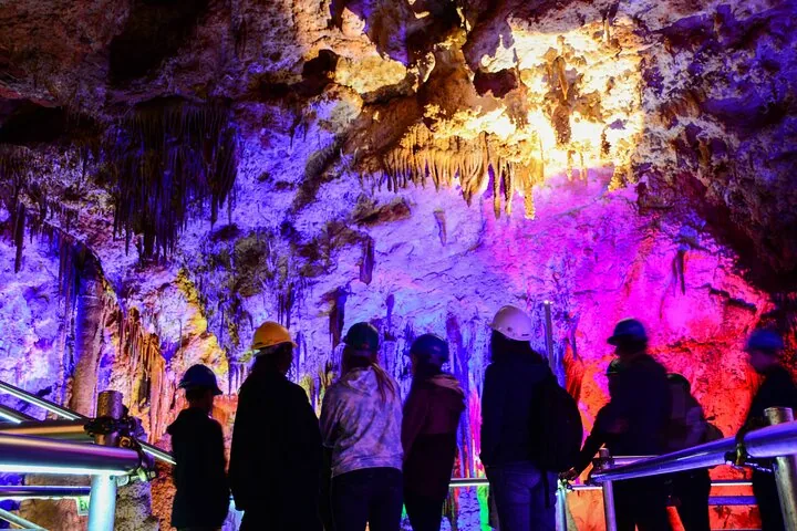 Gruppe von Touristen mit Schutzhelmen erkundet eine bunt beleuchtete Höhle.